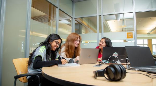Three students sit at a table and look at a laptop screen together