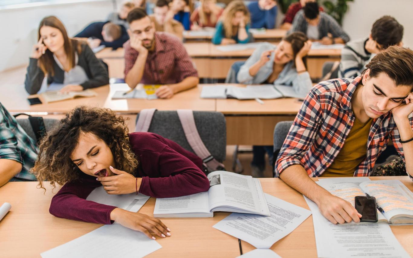 a student on the left yawns while the student next to her has his hand on his face while in a classroom with lots of students in the background